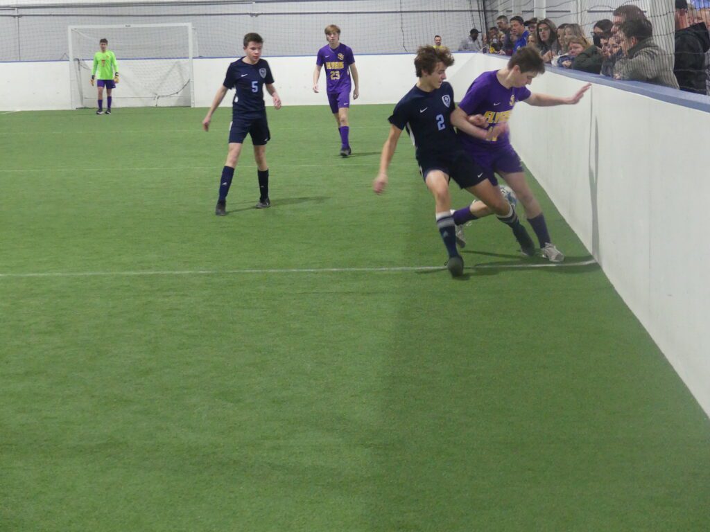 two indoor soccer players of high school age battle for a ball against the boards